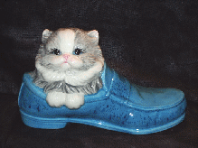 A gray/white, glazed, ceramic cat in a blue-multicolor, glazed, loafer shoe.