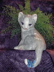 A gray/white glazed, ceramic shelf cat with butterfly on his tail and air fern.