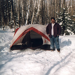 photo- Tim Burns winter camping in the White Mountains of New Hampshire. He is standing next to his tent in the snow.